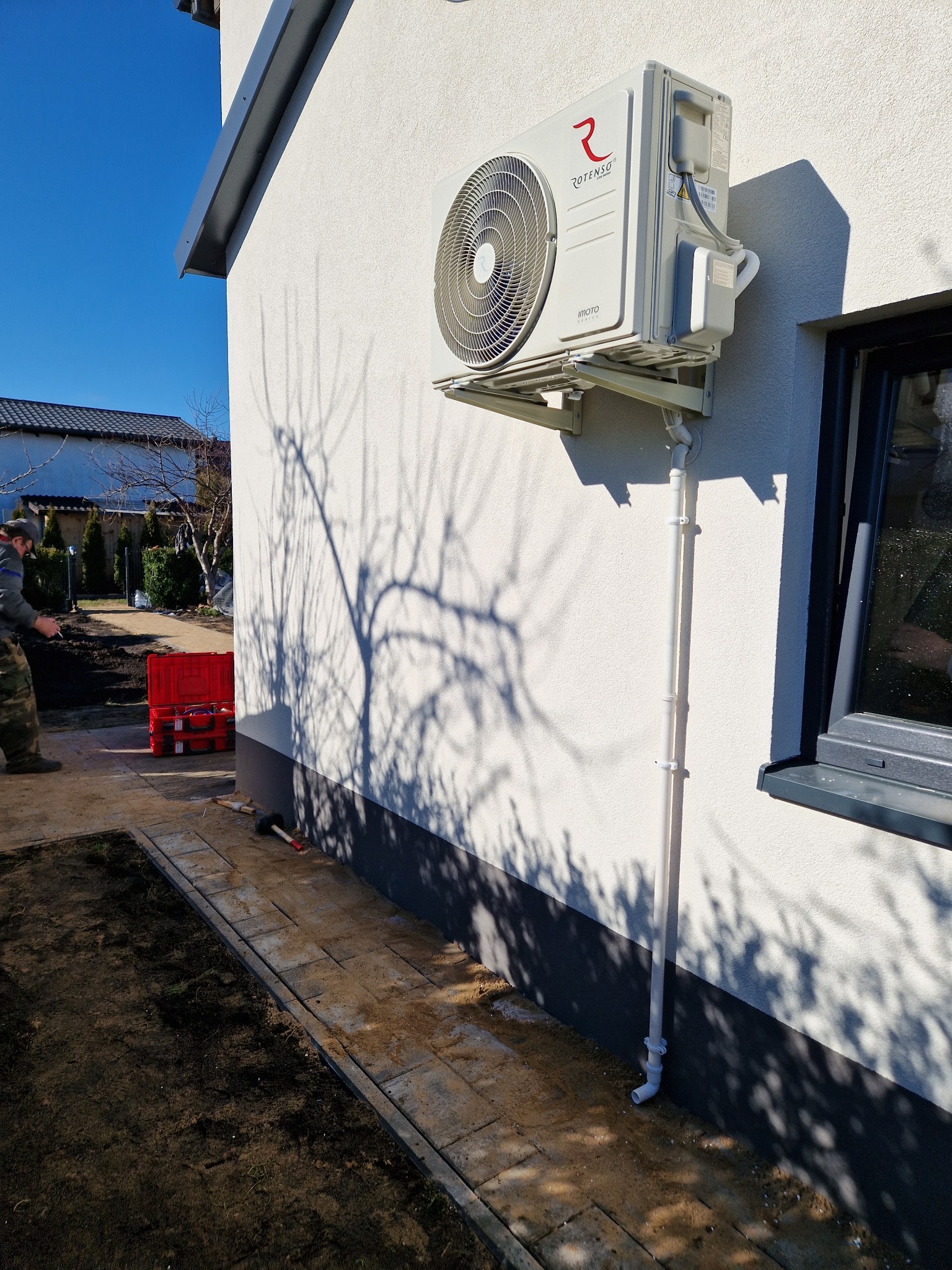 Zewnętrzna jednostka klimatyzacji Rotenso Imoto Series zamontowana na elewacji domu, widoczna rura odprowadzająca skropliny, obok okno, na pierwszym planie częściowo widoczny instalator i czerwona...