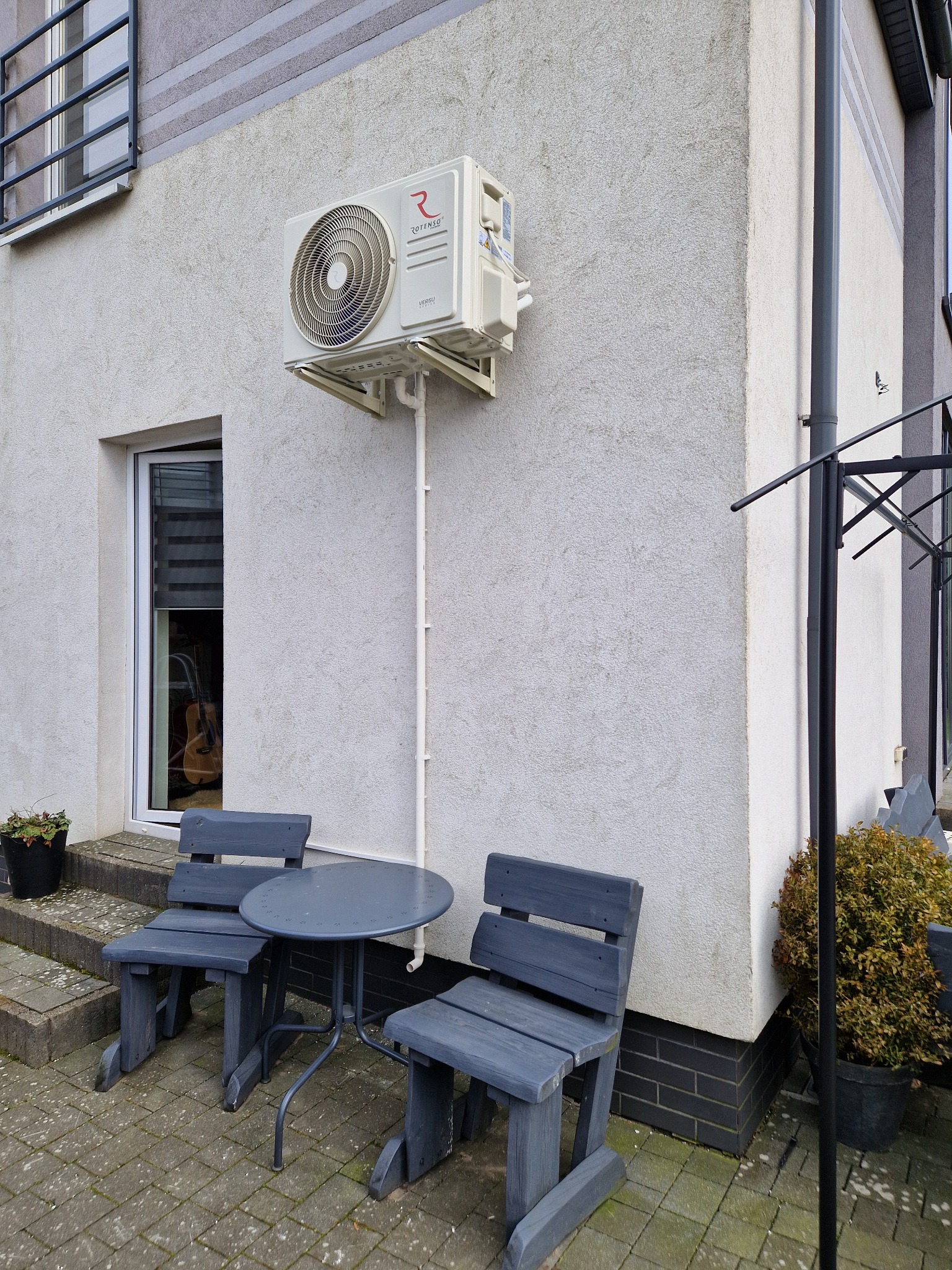 Zewnętrzna jednostka klimatyzacji Rotenso zamontowana na elewacji domu, z widoczną instalacją odprowadzającą skropliny, poniżej stolik i krzesła ogrodowe.