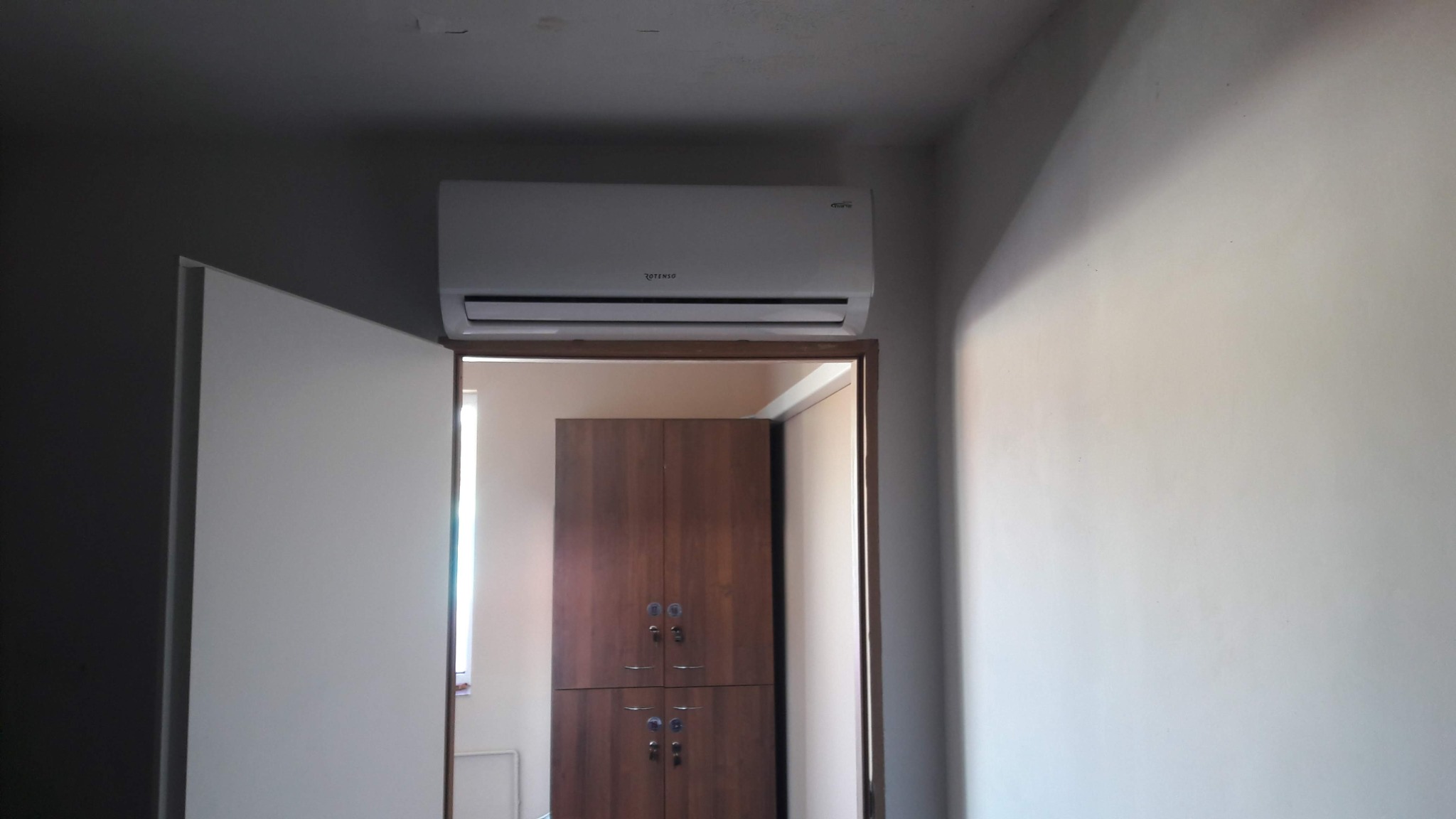 Biała klimatyzacja Rotenso zamontowana nad framugą drzwi w pokoju z widoczną szafą w tle.