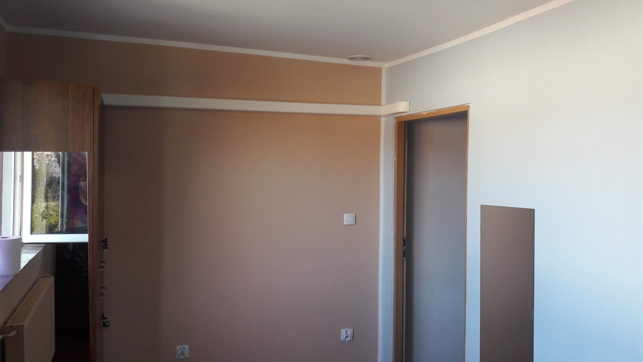 Pomalowane na beżowo ściany pokoju z widoczną instalacją pod klimatyzację w postaci białej listwy na ścianie, oraz zamontowanym panelem ściennym w kolorze szarym na ścianie pomalowanej na jasny...