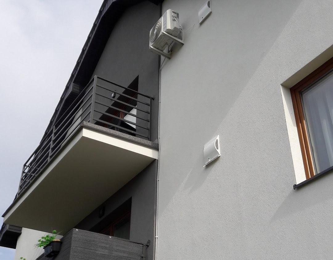 Zewnętrzna jednostka klimatyzacji zamontowana na elewacji dwukondygnacyjnego domu z balkonem, widoczne także nawiewniki ścienne.