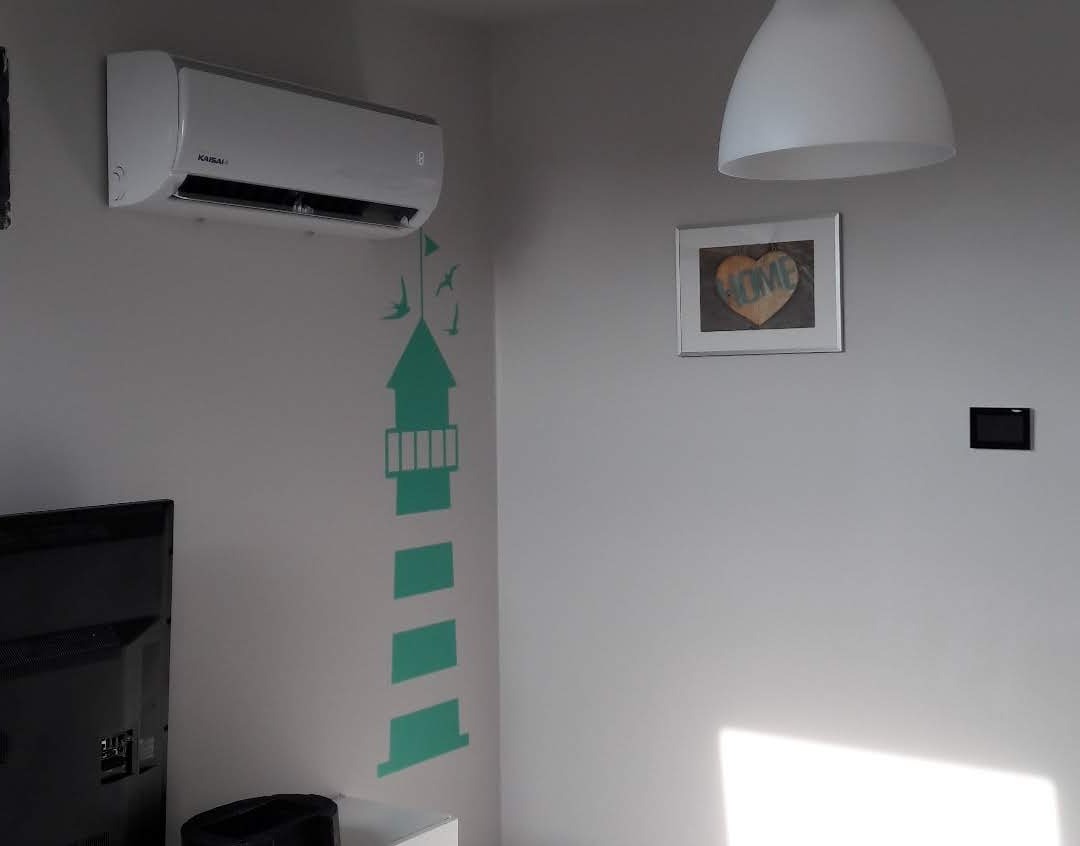 Biały klimatyzator Kaisai zamontowany na szarej ścianie w pokoju z naklejką w kształcie latarni morskiej, lampą i obrazkiem 'HOME'.