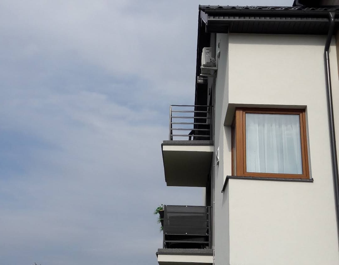 Zewnętrzna jednostka klimatyzacji zamontowana na ścianie budynku z balkonami, widoczna z dołu, na tle pochmurnego nieba.