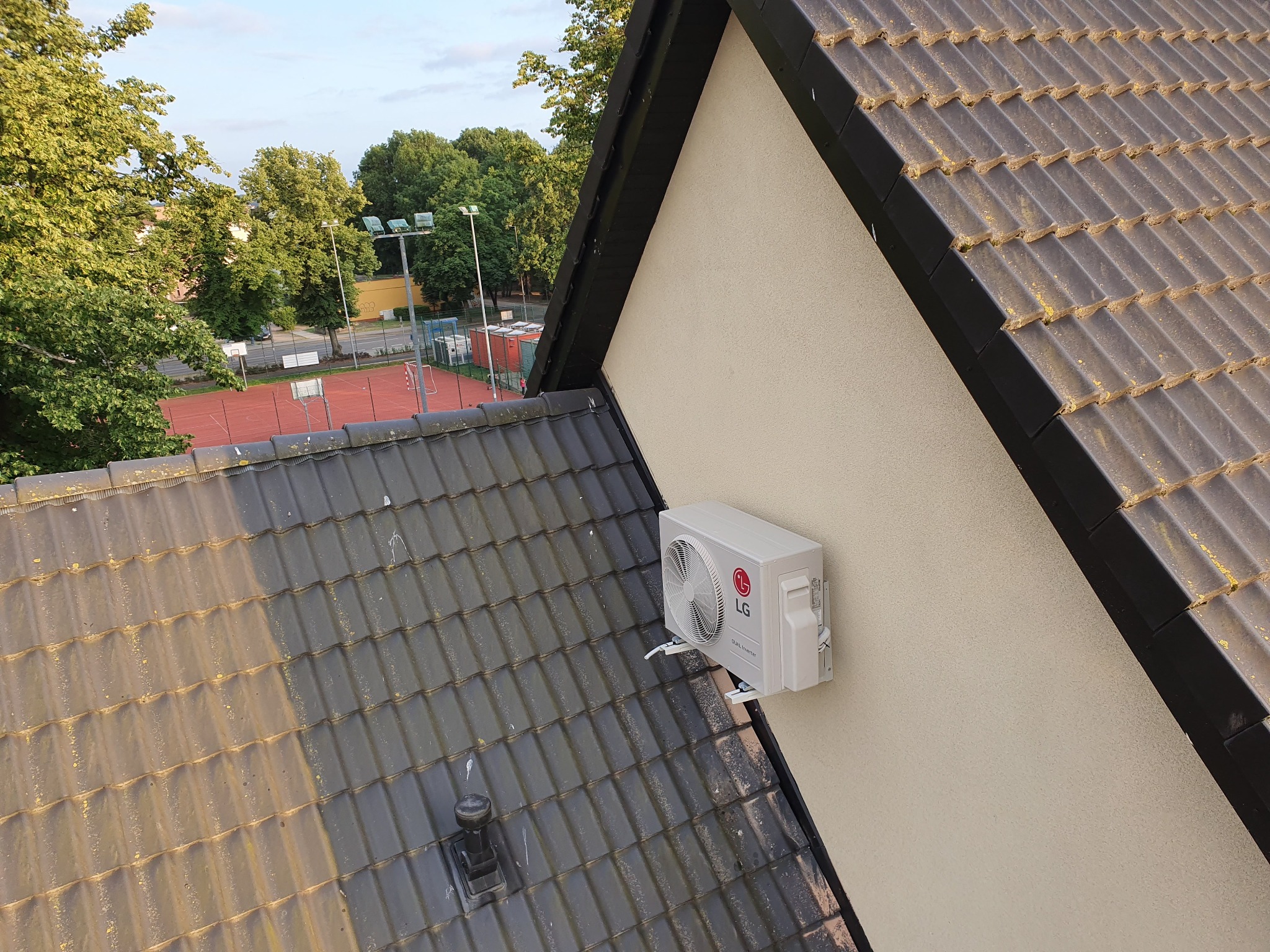 Zewnętrzna jednostka klimatyzacji LG zamontowana na ścianie domu, widok z góry z fragmentem dachu pokrytego brązową dachówką, w tle drzewa i boisko sportowe.