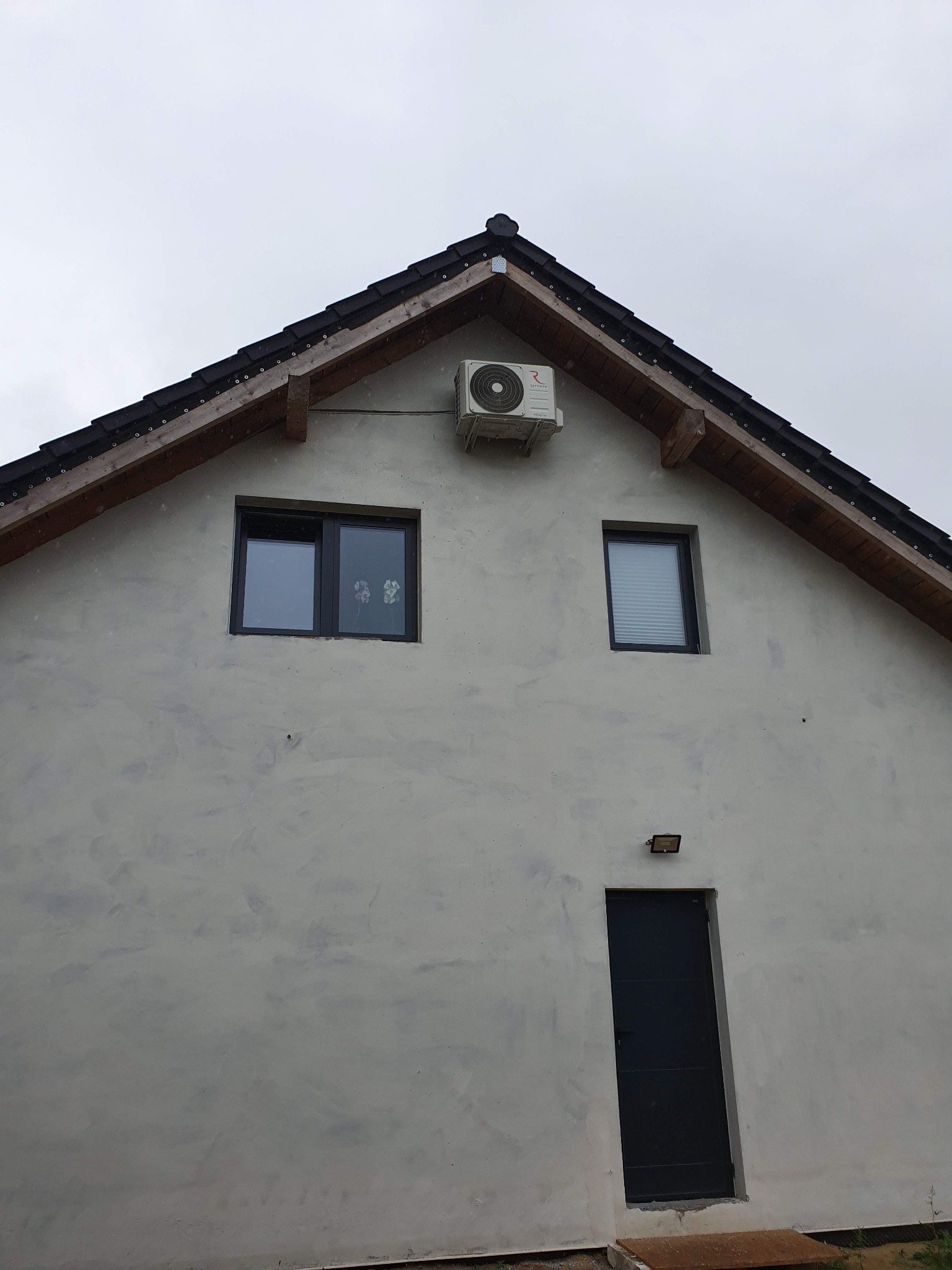Zewnętrzna jednostka klimatyzacji zamontowana na szczycie ściany szczytowej domu jednorodzinnego z dwoma oknami i ciemnymi drzwiami wejściowymi.