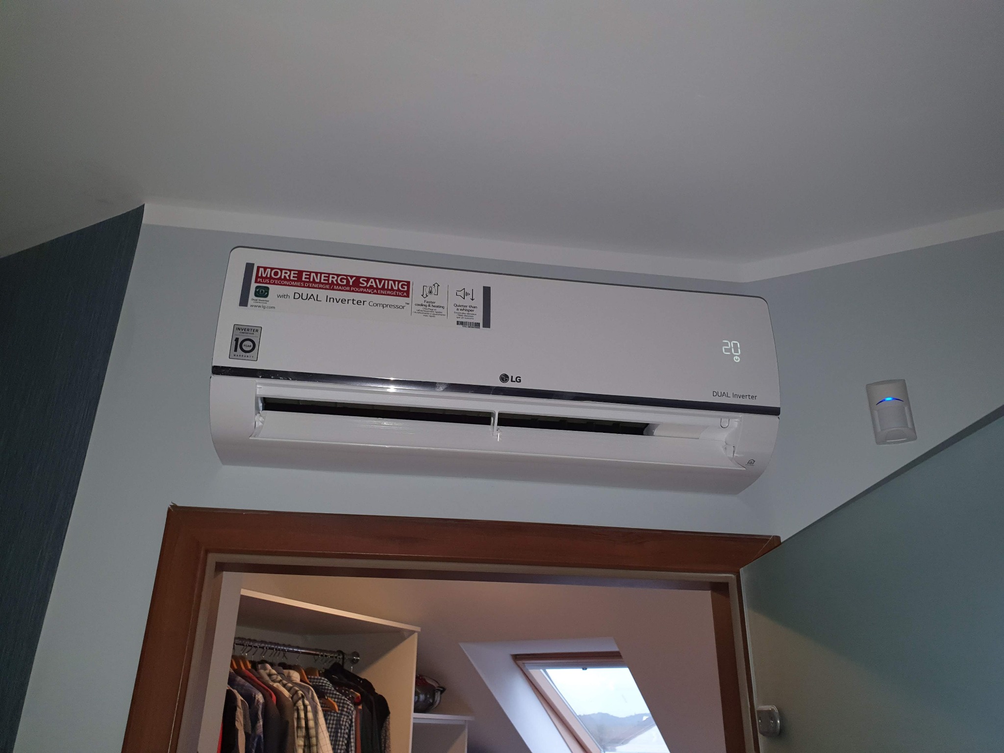 Biała jednostka wewnętrzna klimatyzatora LG Dual Inverter zamontowana nad drzwiami garderoby w jasnym pomieszczeniu. Widoczny czujnik ruchu na ścianie obok.