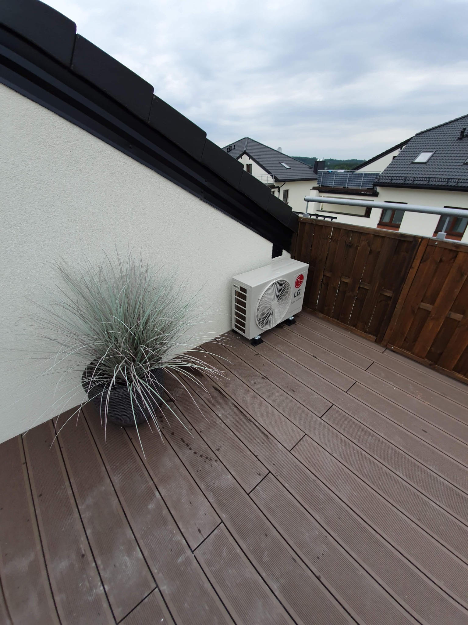 Zewnętrzna jednostka klimatyzacji LG Dual Inverter na drewnianym tarasie, obok donica z ozdobną trawą, w tle domy jednorodzinne pod pochmurnym niebem.