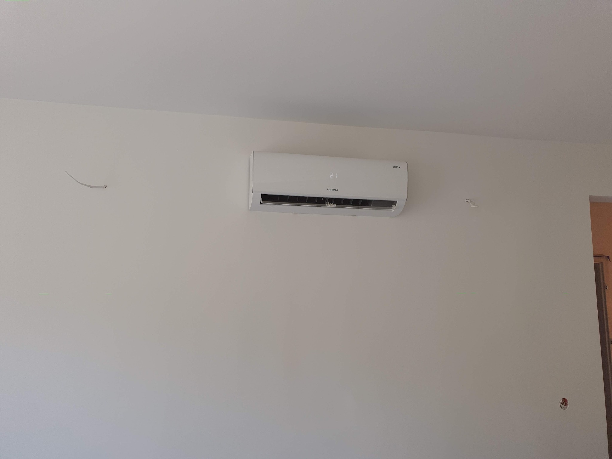 Biały klimatyzator Rotenso zamontowany na jasnej ścianie w pomieszczeniu, wyświetlający temperaturę 21 stopni Celsjusza.