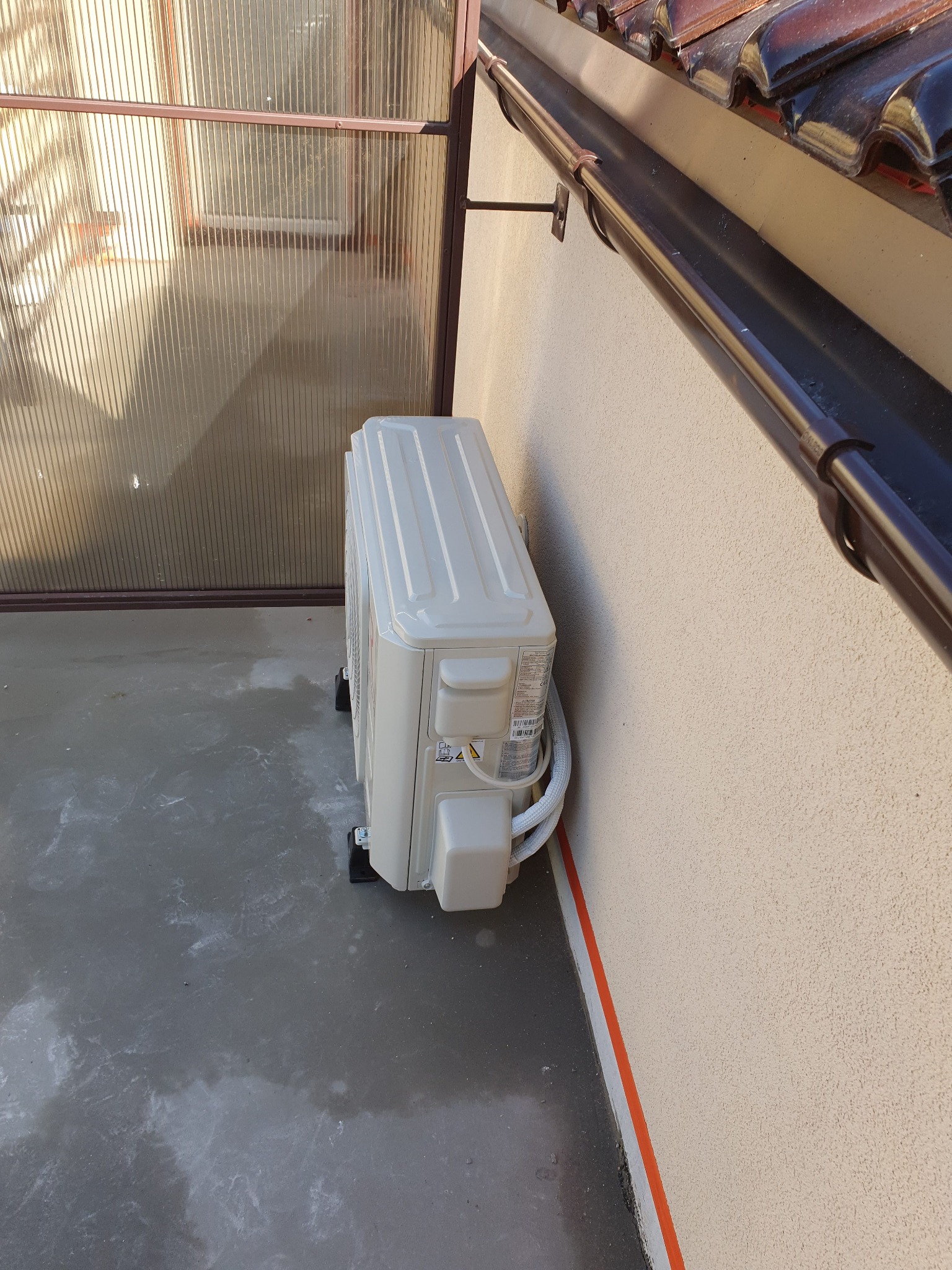 Zewnętrzna jednostka klimatyzacji zamontowana przy ścianie budynku z widoczną rynną i elementami elewacji. Białe przewody prowadzą do wnętrza. Podłoże betonowe z plamami.