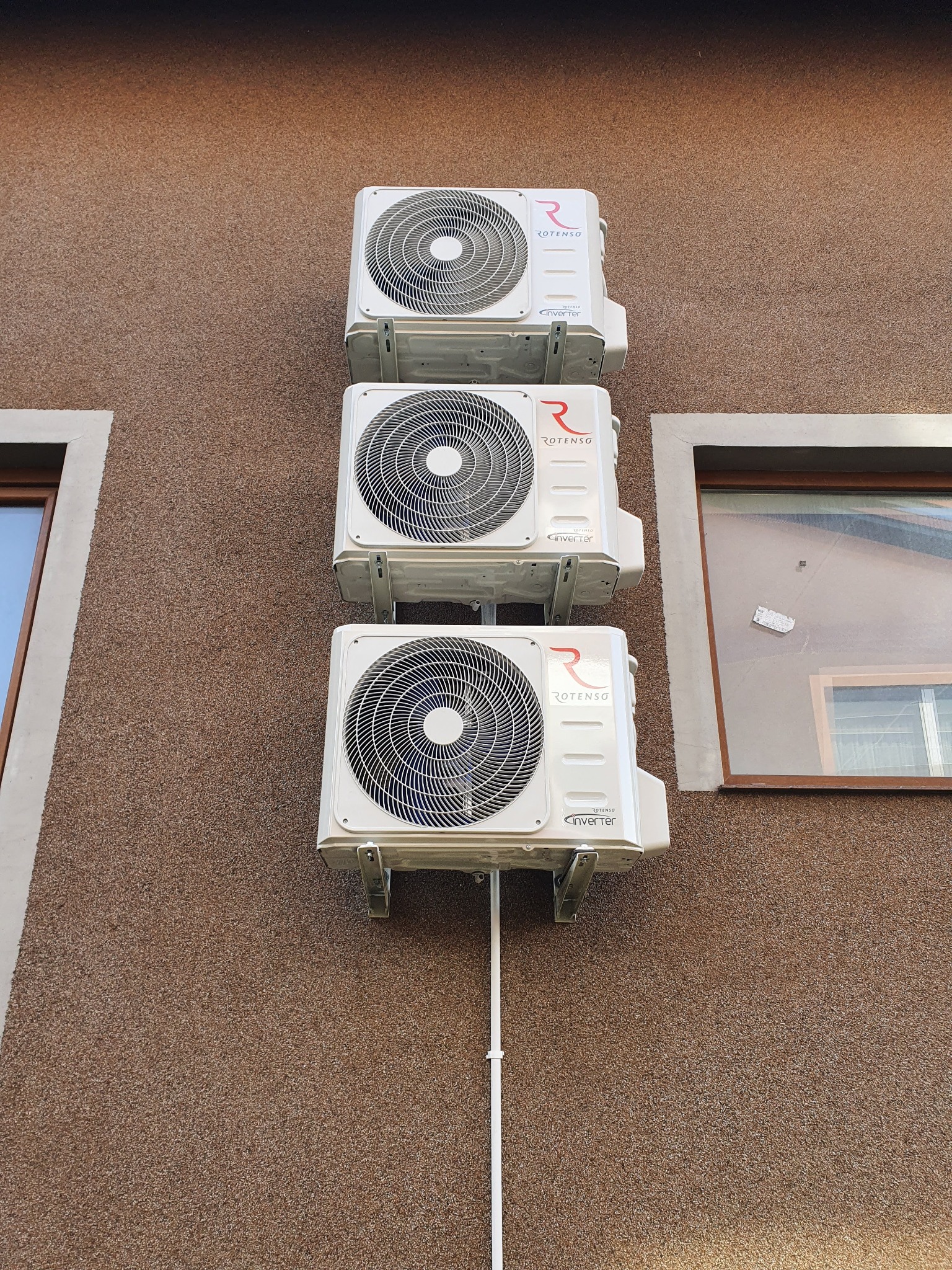 Trzy białe jednostki zewnętrzne klimatyzacji Rotenso Inverter zamontowane pionowo na brązowej elewacji budynku, widoczne okna z białymi ramami po bokach.