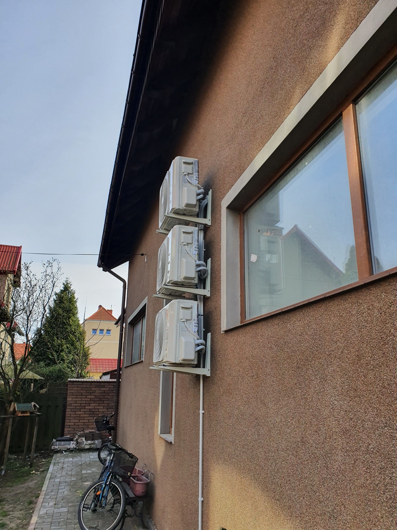 Trzy jednostki zewnętrzne klimatyzacji zamontowane pionowo na ścianie domu z oknem, widoczne rowery i otoczenie budynku.