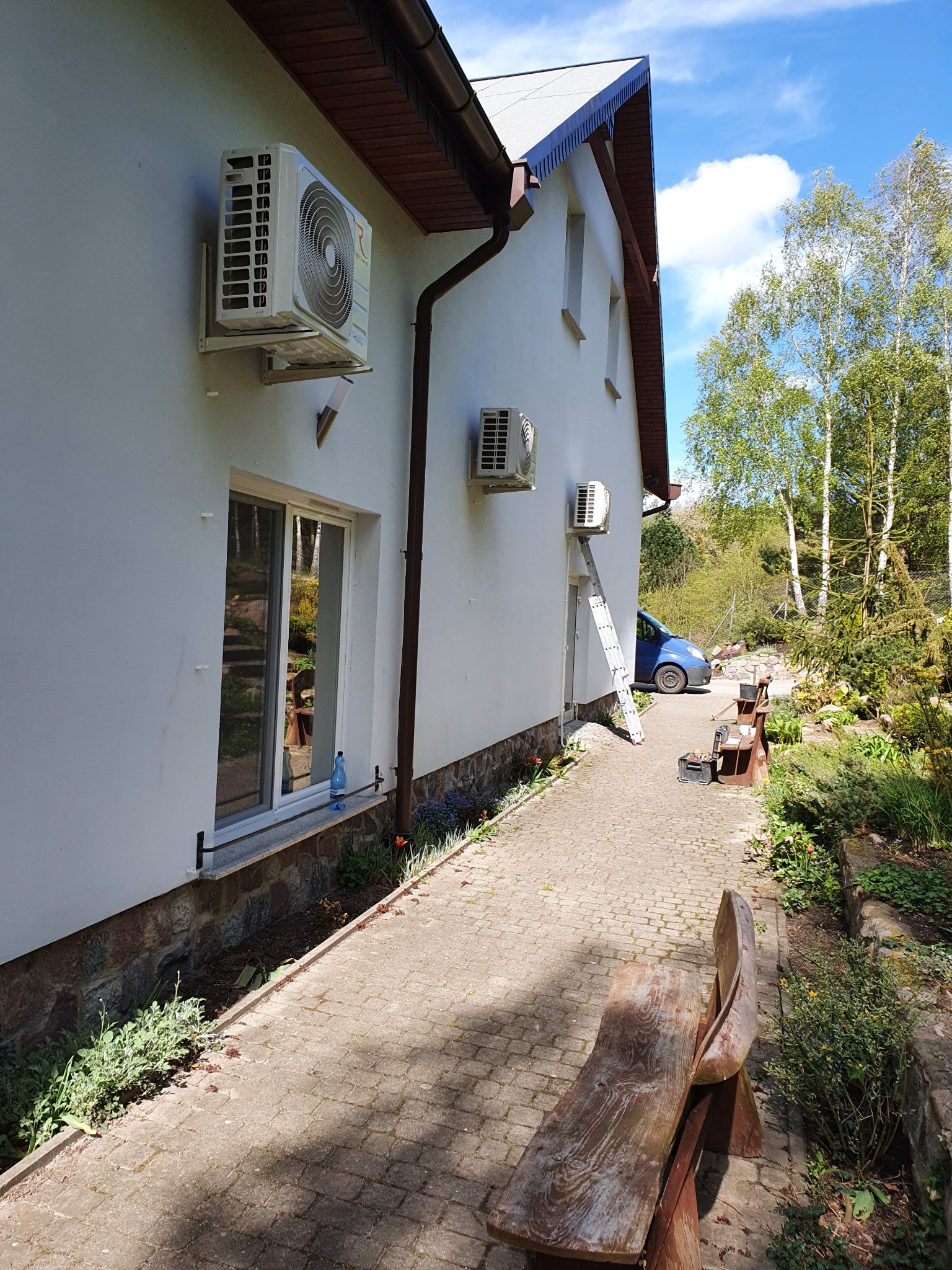 Dwa klimatyzatory zamontowane na ścianie domu w Lęborku. Na chodniku oparta drabina, obok zaparkowany niebieski samochód, w tle drzewa.