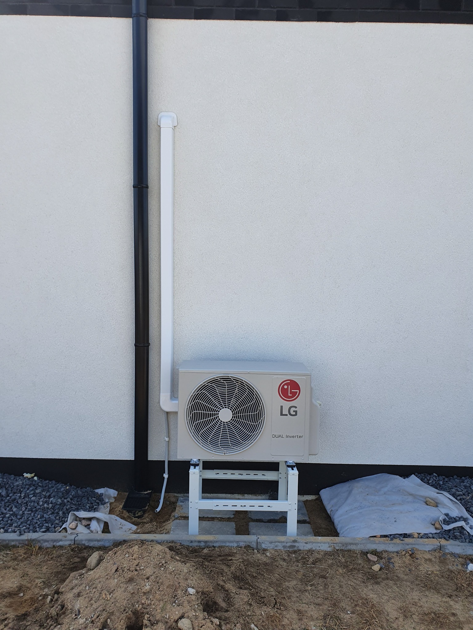 Zewnętrzna jednostka klimatyzacji LG Dual Inverter zamontowana na metalowym stojaku przy białej ścianie budynku z czarną rynną, widoczny fragment terenu z kamieniami i ziemią.