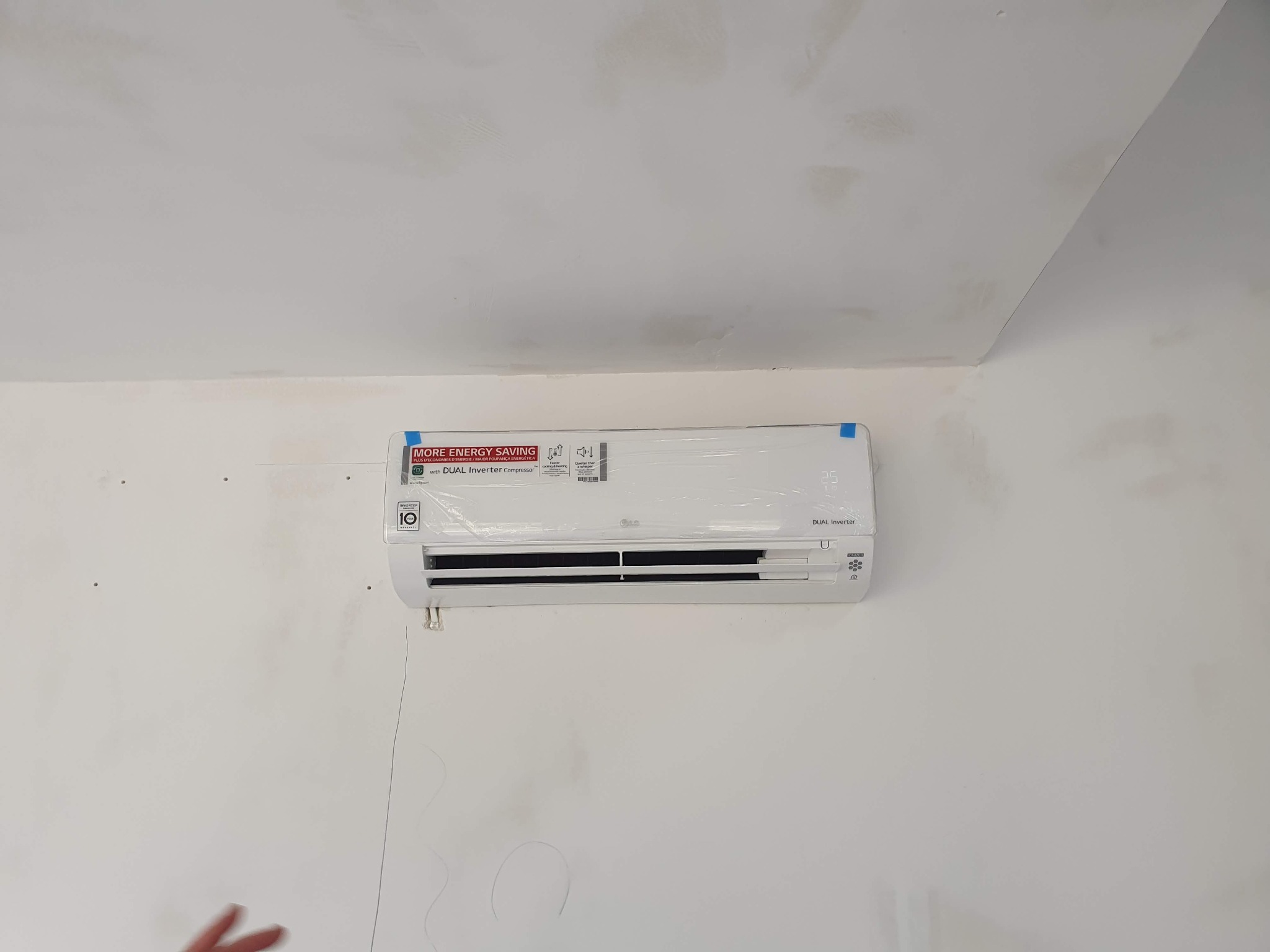 Biała jednostka wewnętrzna klimatyzacji LG Dual Inverter, jeszcze w folii ochronnej, zamontowana na białej ścianie z widocznymi śladami po wierceniu i niedoskonałościami wykończenia.