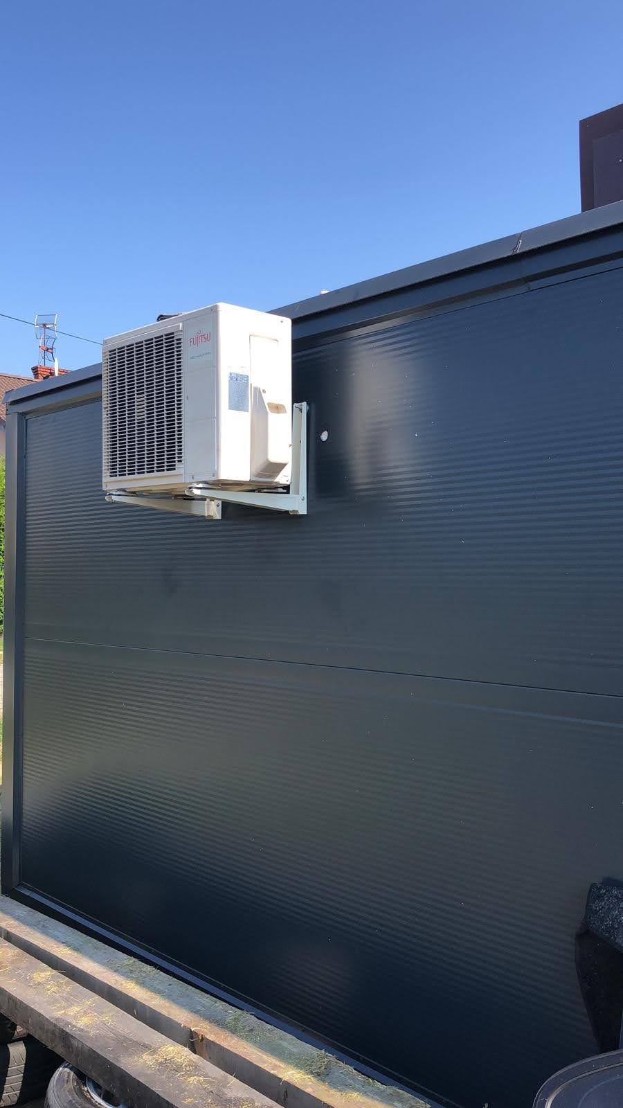 Biała jednostka zewnętrzna klimatyzatora marki Fujitsu zamontowana na ciemnoszarej, blaszanej ścianie budynku z widocznymi elementami dachu w tle.