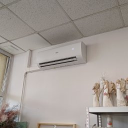 ARKEN - Biały klimatyzator Haier zamontowany na ścianie w kwiaciarni, widoczne dekoracyjne figurki aniołów na półce obok.