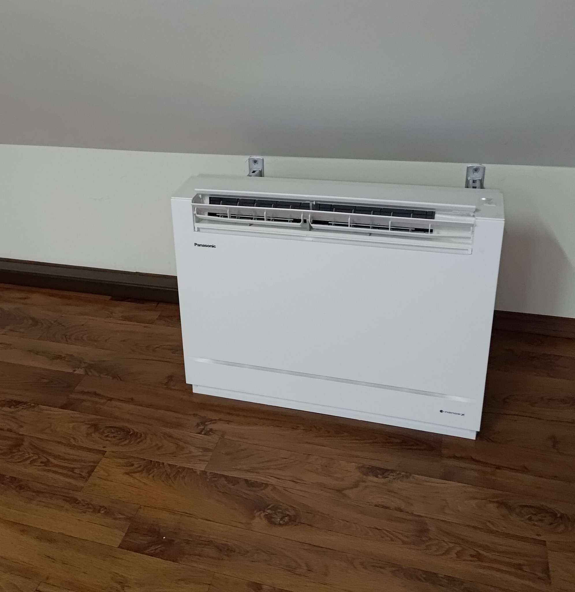 Biały klimatyzator konsolowy Panasonic zamontowany na ścianie kolankowej poddasza za pomocą dystansów, widoczny na tle drewnianej podłogi i białej ściany.