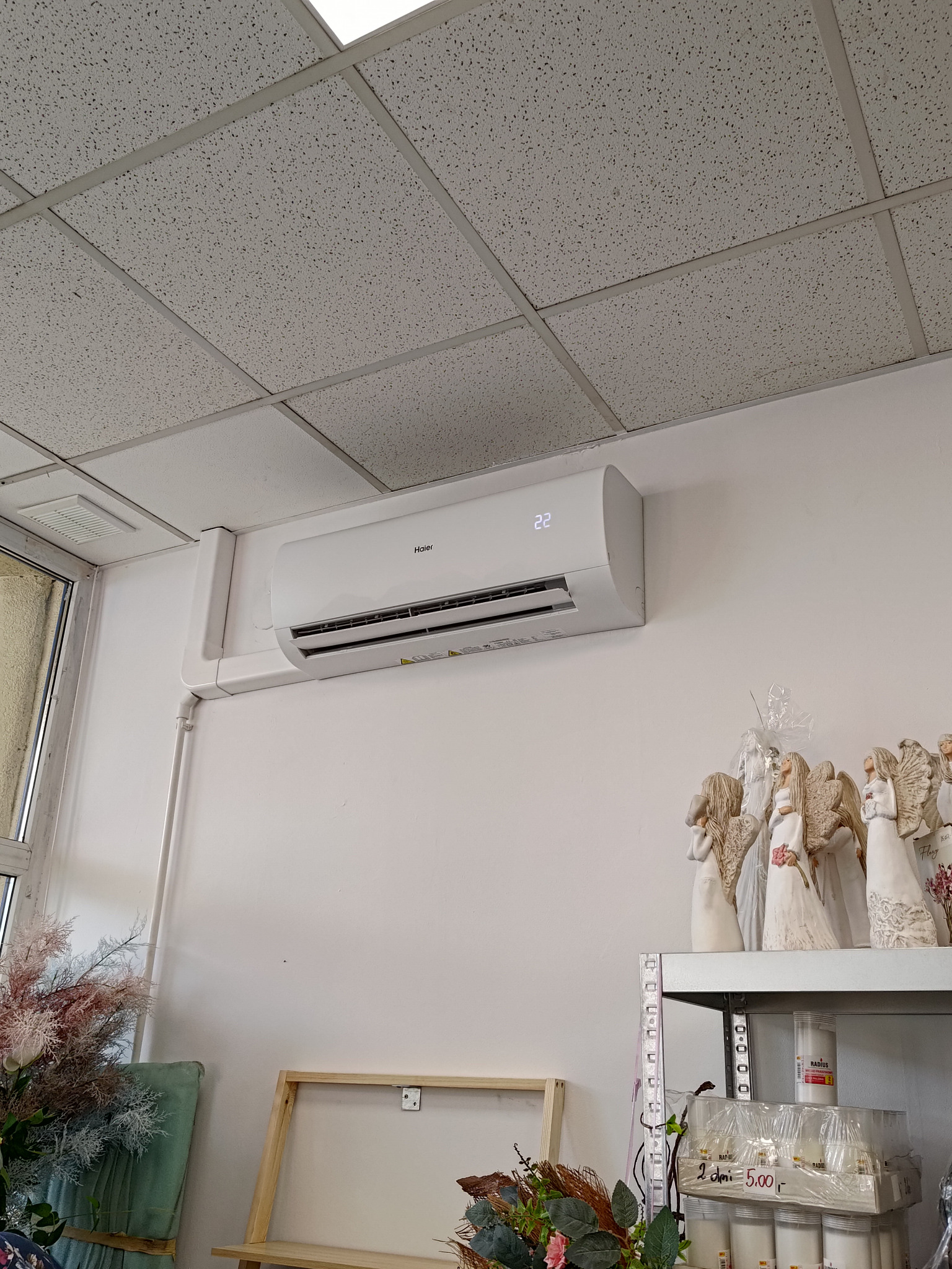 Biały klimatyzator Haier zamontowany na ścianie w kwiaciarni, widoczne dekoracyjne figurki aniołów na półce obok.