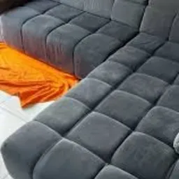 Szara, modułowa kanapa narożna z kwadratowymi siedziskami na białej podłodze, zabezpieczona pomarańczową folią malarską.