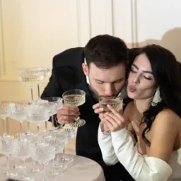 Elegancka para w strojach wieczorowych pije szampana z piramidy kieliszk&oacute;w, kobieta z ustami ułożonymi w pocałunek, mężczyzna w garniturze, subtelne oświetlenie.