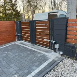 Nowoczesna brama przesuwna z automatyką, drewniane wypełnienie z poziomych listew, szara kostka brukowa, widoczny napęd i skrzynka sterująca, w tle biały samochód dostawczy.