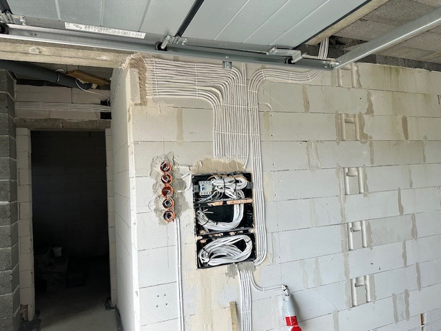 Świeżo ułożone przewody elektryczne w białych rurkach na ścianie z betonu komórkowego, widoczna skrzynka rozdzielcza z przewodami oraz puszki instalacyjne pod gniazdka, szyny do bramy garażowej...