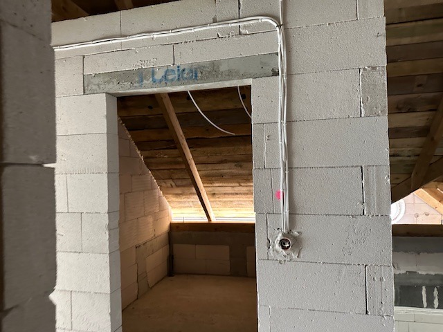 Surowy stan poddasza z białych bloczków, widoczne kable elektryczne poprowadzone po ścianie, puszka elektryczna zamontowana w ścianie, drewniana konstrukcja dachu.