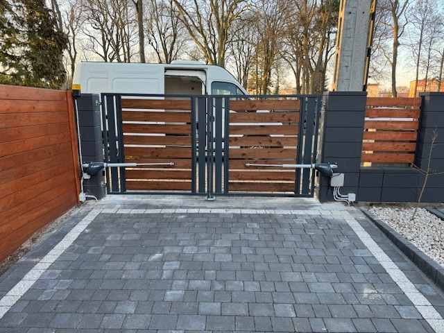 Nowoczesna brama dwuskrzydłowa z automatyką, widoczne siłowniki, drewniane wypełnienie, szara kostka brukowa na podjeździe, biały bus w tle.