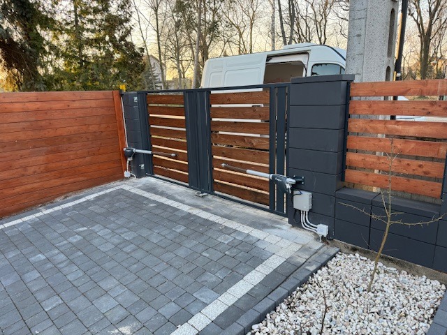 Nowoczesna brama przesuwna z automatyką, drewniane wypełnienie z poziomych listew, szara kostka brukowa, widoczny napęd i skrzynka sterująca, w tle biały samochód dostawczy.