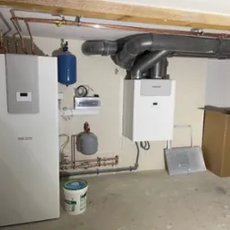 Dwie jednostki grzewcze marki Stiebel Eltron, jedna stojąca i jedna wisząca, z widocznymi rurami miedzianymi i przewodami wentylacyjnymi w pomieszczeniu technicznym.