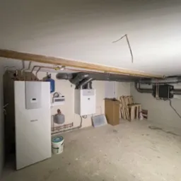 Instalacja urządzeń grzewczych i wentylacyjnych w surowym pomieszczeniu: dwa kotły, zbiornik wyrównawczy, system rur wentylacyjnych pod sufitem, widoczne elementy montażowe.