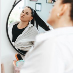 Studio Inna Koteliukh - Kobieta z czarnymi włosami spiętymi w wysoki kucyk, ubrana w białą koszulę, przegląda się w okrągłym lustrze w czarnej ramie, obok widoczne szczotki do włosów w ozdobnym pudełku.