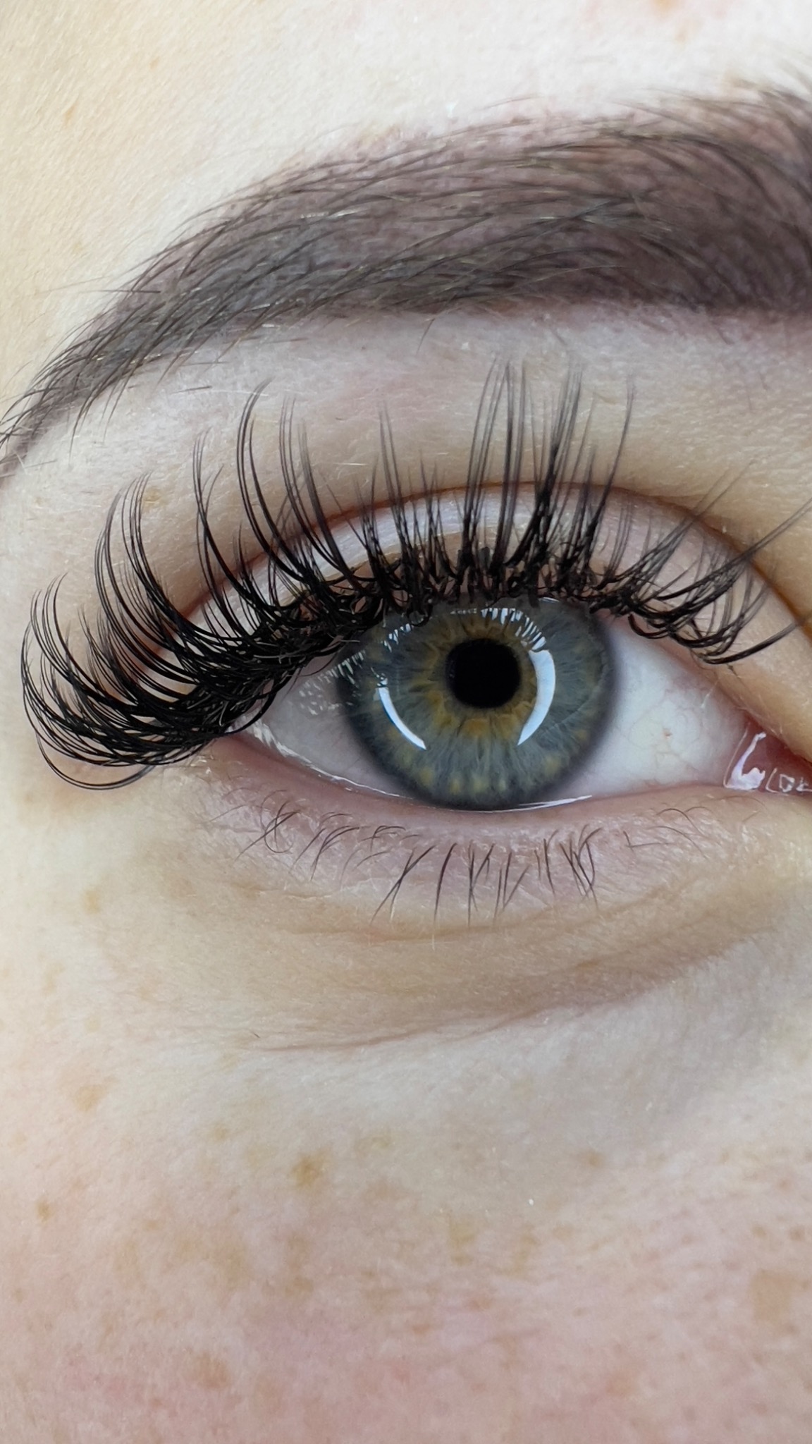 Zbliżenie na oko z wyraźnie przedłużonymi i zagęszczonymi czarnymi rzęsami, widoczna tęczówka w odcieniach szarości i zieleni oraz naturalny makijaż brwi.