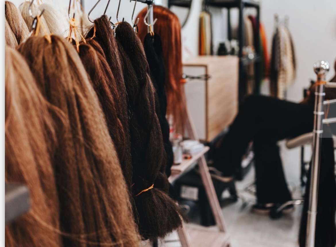 Wiązki włosów syntetycznych w różnych odcieniach brązu, blondu i czerni, zawieszone na metalowym wieszaku w salonie, z widocznym stanowiskiem pracy w tle.