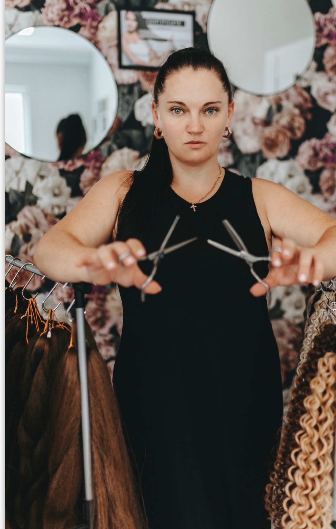 Kobieta z poważnym wyrazem twarzy trzymająca dwie pary nożyczek fryzjerskich na tle kwiecistej tapety, lustra i wieszaka z doczepianymi włosami w różnych kolorach i teksturach.