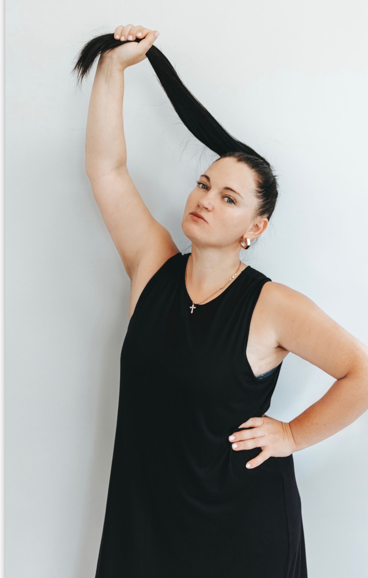 Kobieta z ciemnymi włosami uniesionymi do góry, ubrana w czarną sukienkę, pozuje na tle jasnej ściany, prezentując długość i kondycję włosów.