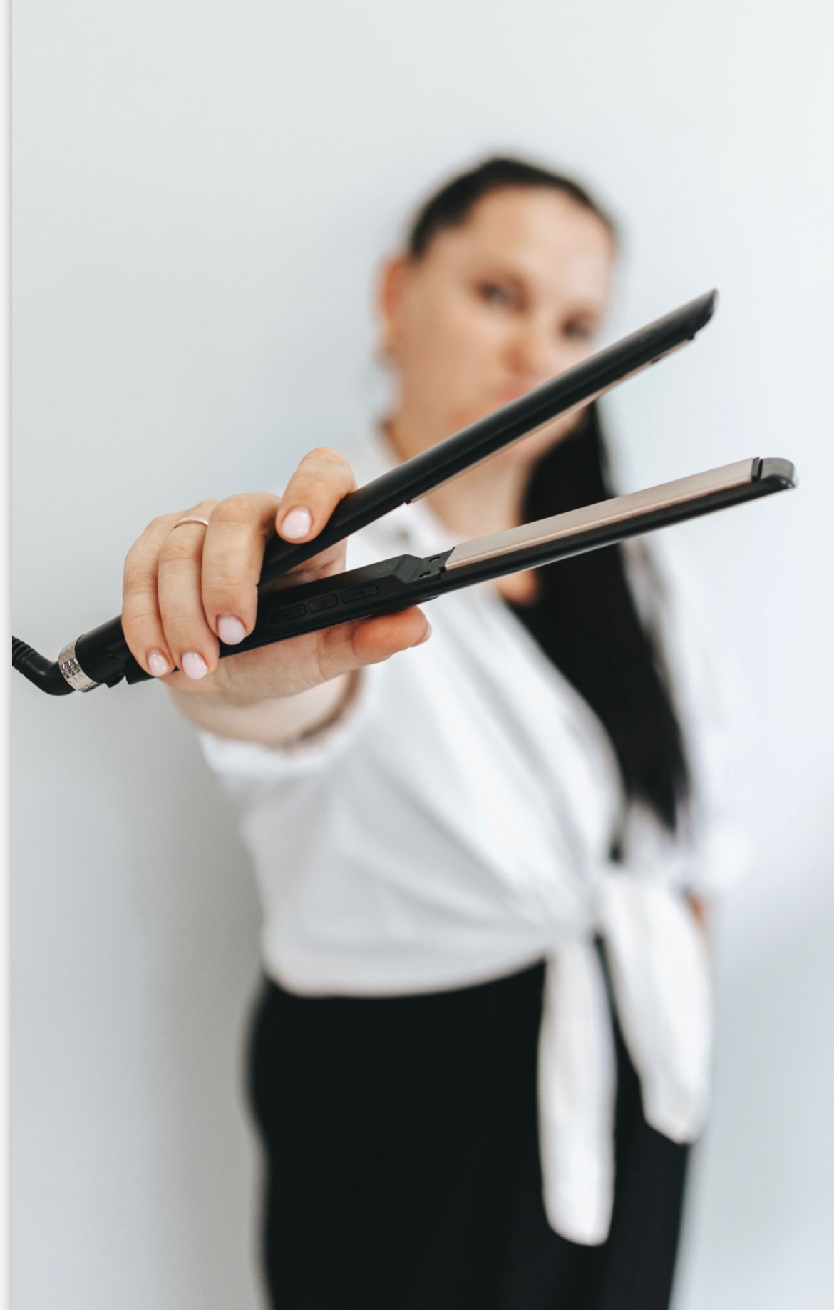 Kobieta w białej koszuli prezentuje prostownicę do włosów z ceramicznymi płytkami, trzymając ją w dłoni z pierścionkiem.
