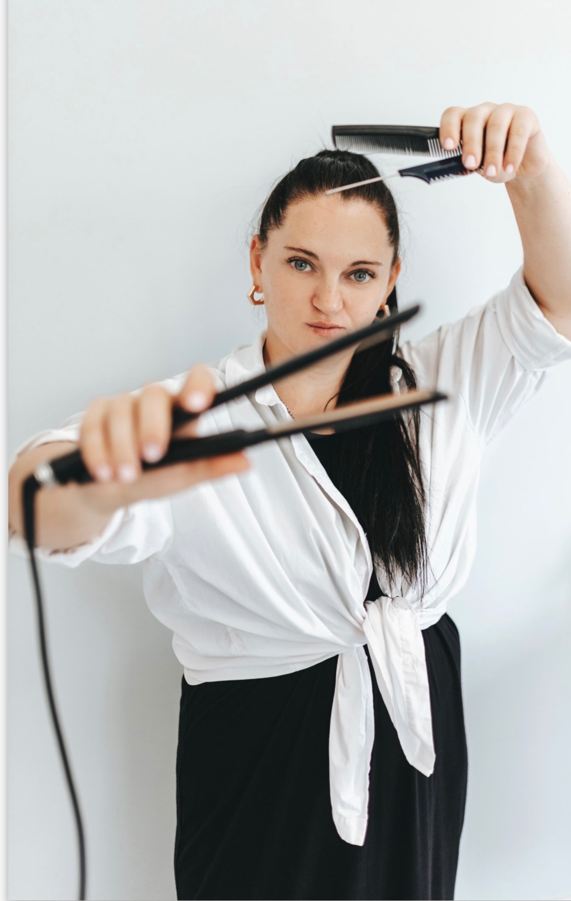 Kobieta z ciemnymi włosami, w białej koszuli, trzyma prostownicę i grzebień przygotowując się do stylizacji włosów.