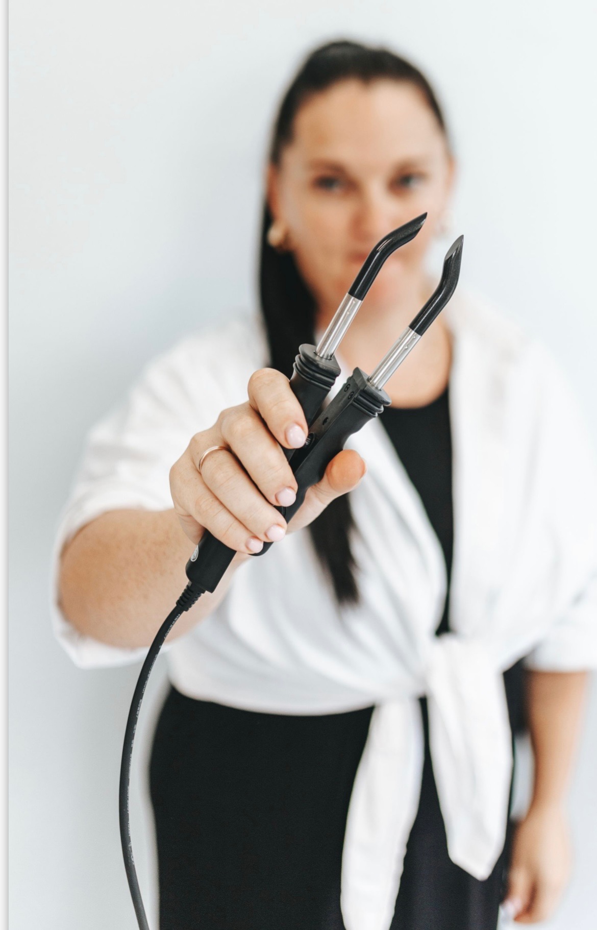 Kobieta w białej bluzce trzyma w dłoni urządzenie do stylizacji włosów z dwoma czarnymi końcówkami, na palcu ma złoty pierścionek.