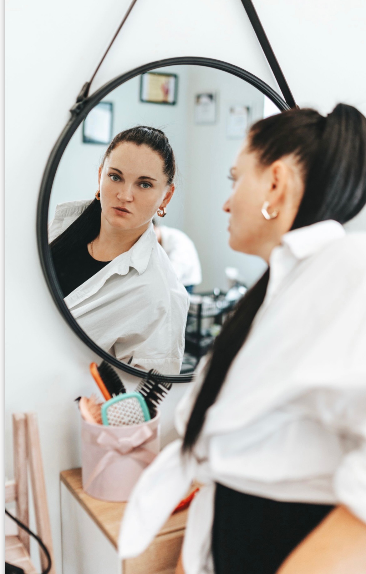 Kobieta z ciemnymi włosami spiętymi w wysoki kucyk, ubrana w białą koszulę, patrzy w okrągłe lustro, w tle widoczne akcesoria do włosów w różowym pudełku.