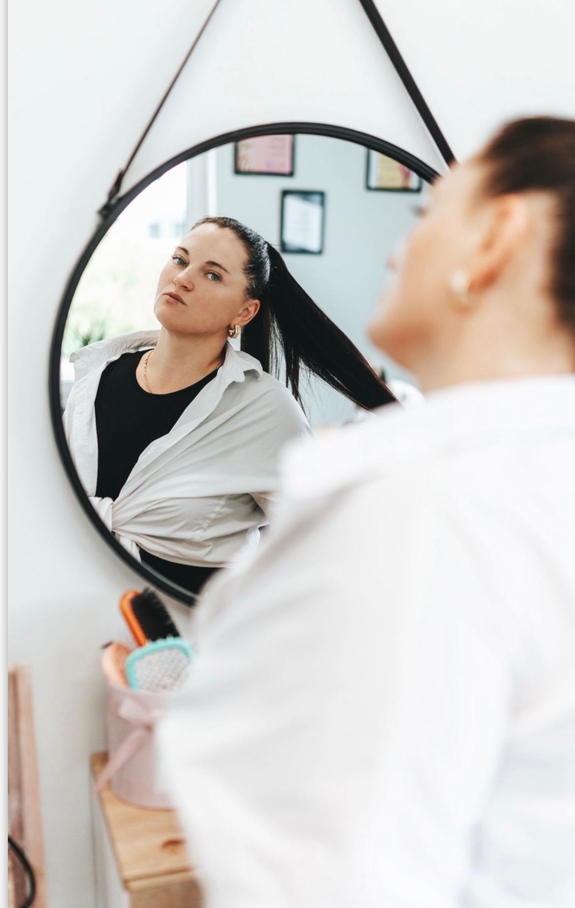 Kobieta z czarnymi włosami spiętymi w wysoki kucyk, ubrana w białą koszulę, przegląda się w okrągłym lustrze w czarnej ramie, obok widoczne szczotki do włosów w ozdobnym pudełku.