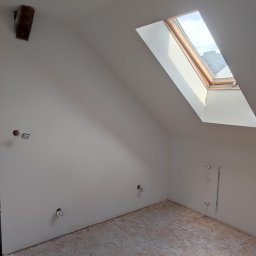 Uloo-instal - Poddasze po szpachlowaniu ścian i przygotowaniu podłogi z płyt OSB, z oknem dachowym i wystającymi kablami elektrycznymi i rurami hydraulicznymi, surowa żarówka zwisa z sufitu.