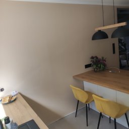 Uloo-instal - Beżowa ściana w kuchni z drewnianym blatem, czarnym zlewem, żółtymi krzesłami i czarną lampą wiszącą, na blacie wazon z fioletowymi kwiatami i talerz z goframi.