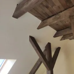 Wyeksponowane drewniane belki stropowe w jasnym, odnowionym wnętrzu poddasza, z widocznym oknem dachowym w lewym dolnym rogu.