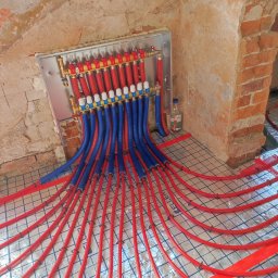 Uloo-instal - Rozdzielacz ogrzewania podłogowego z czerwonymi i niebieskimi rurami na folii aluminiowej, widok z góry, surowa ściana w tle.