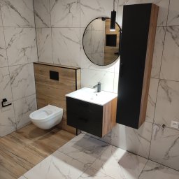 Uloo-instal - Nowoczesna łazienka z marmurowymi płytkami, okrągłym lustrem i wiszącą szafką. Uwagę zwraca kontrast faktur drewna i czerni w zabudowie toalety i umywalki.
