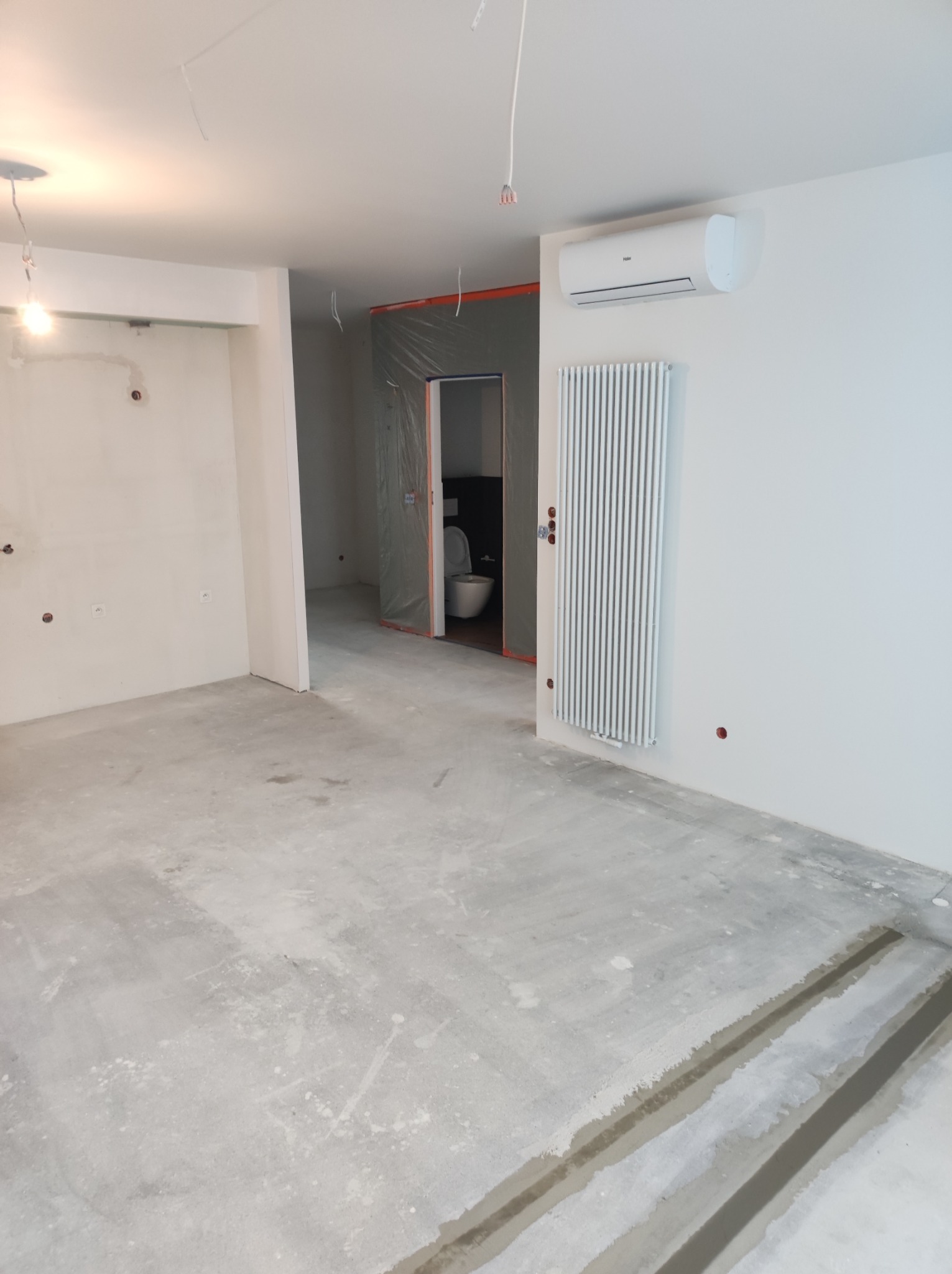 Wnętrze w Poniecu w trakcie prac wykończeniowych, widoczny surowy betonowy podkład, grzejnik na ścianie, klimatyzator i zabezpieczone folią wejście do łazienki.