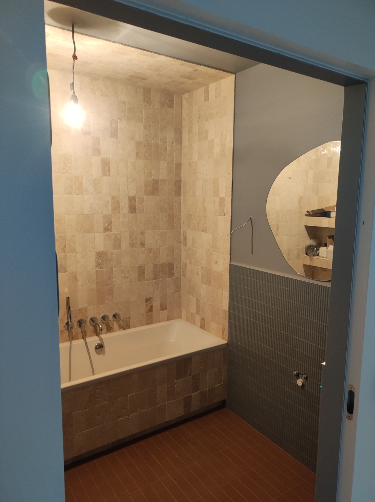 Wykończona łazienka z wanną obłożoną beżowymi płytkami, lustrem o nieregularnym kształcie i brązową podłogą. Wisząca żarówka bez klosza.