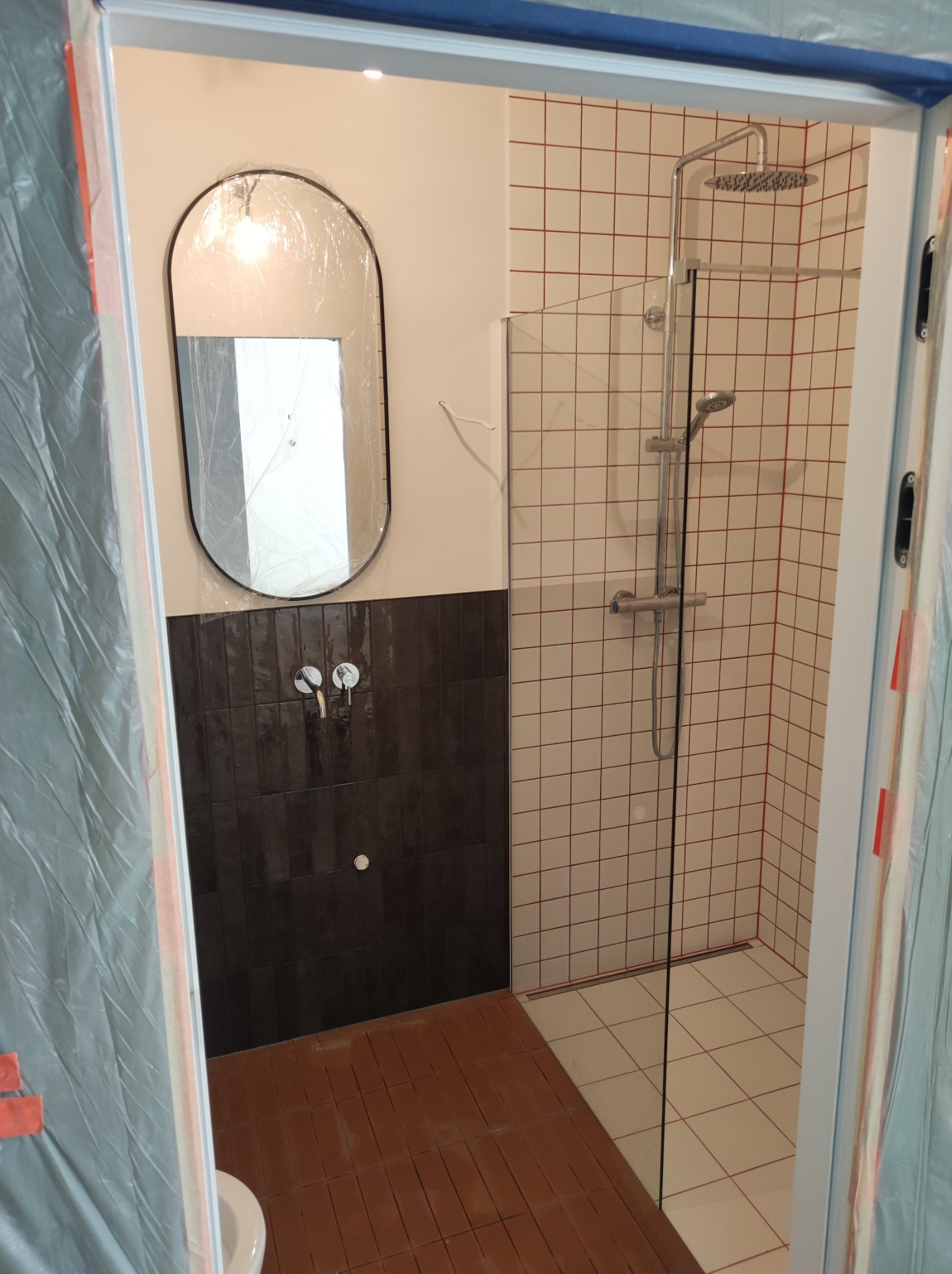 Nowoczesna kabina prysznicowa z przeszkloną ścianką, wykończona białymi płytkami z czerwoną fugą, ciemnobrązowe płytki pod umywalką, owalne lustro z czarną ramą, podłoga z brązowych płytek...