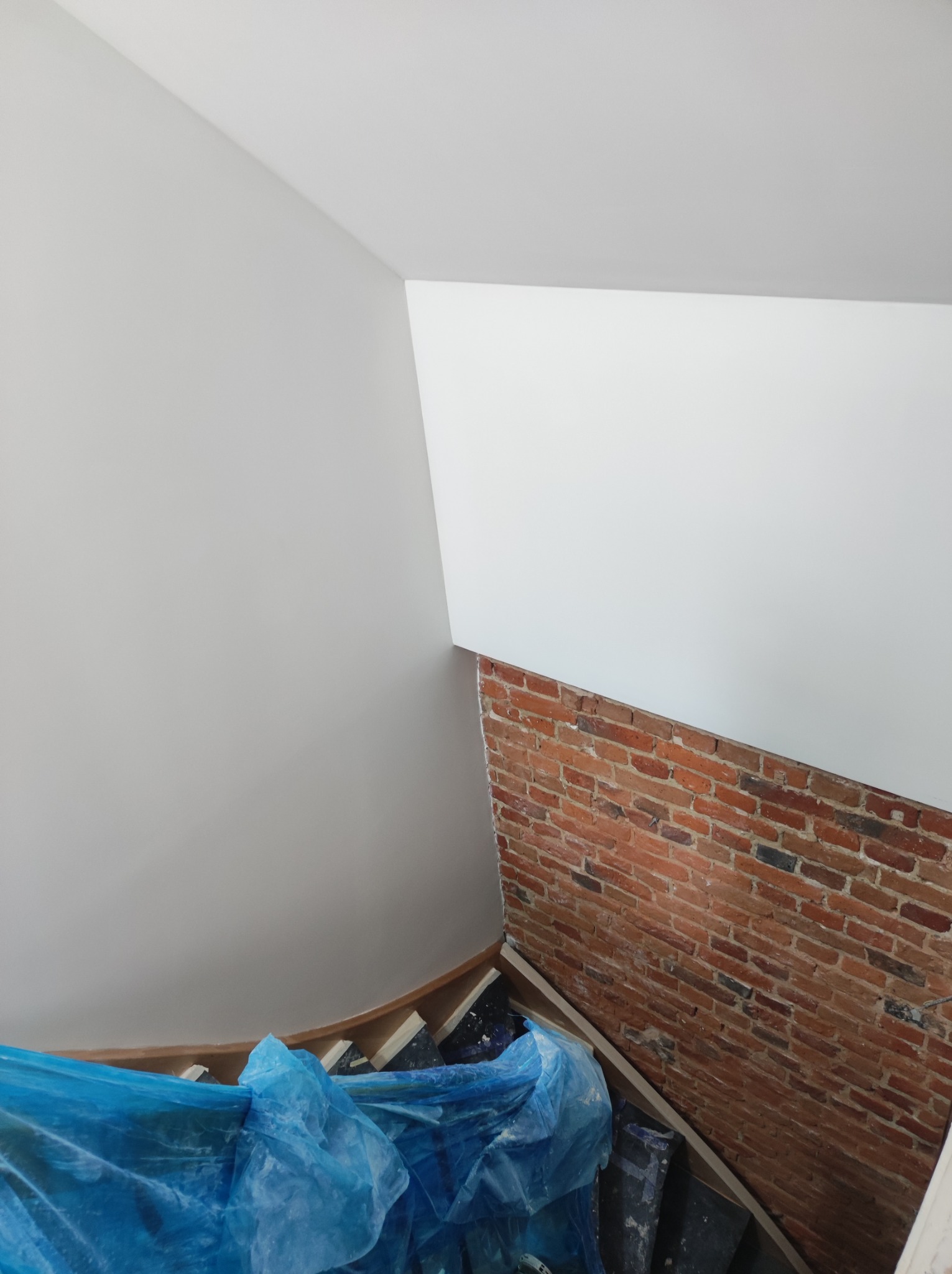 Widok z góry na fragment wnętrza w trakcie prac: pomalowane na szaro ściany, fragment ściany z czerwonej cegły, zabezpieczone folią schody.